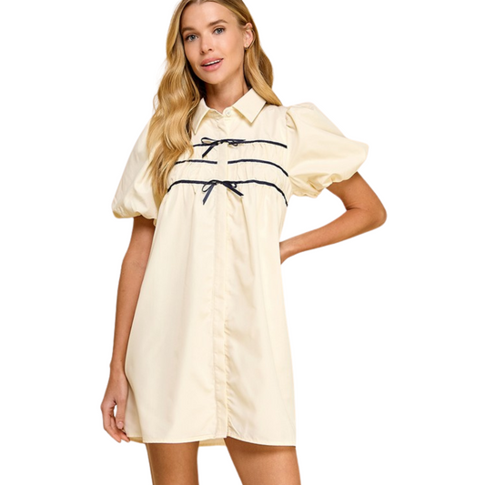 This cream-colored dress features a button-up short sleeve design with elegant contrast ribbon accents and charming bow ties at the center front. Fully lined for comfort and a polished finish. Perfect for a sophisticated and refined look.